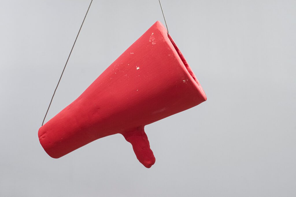 Sculpture by artist Laura Mahnke: Bright red ceramic megaphone hanging from the ceiling in front of a gray wall.