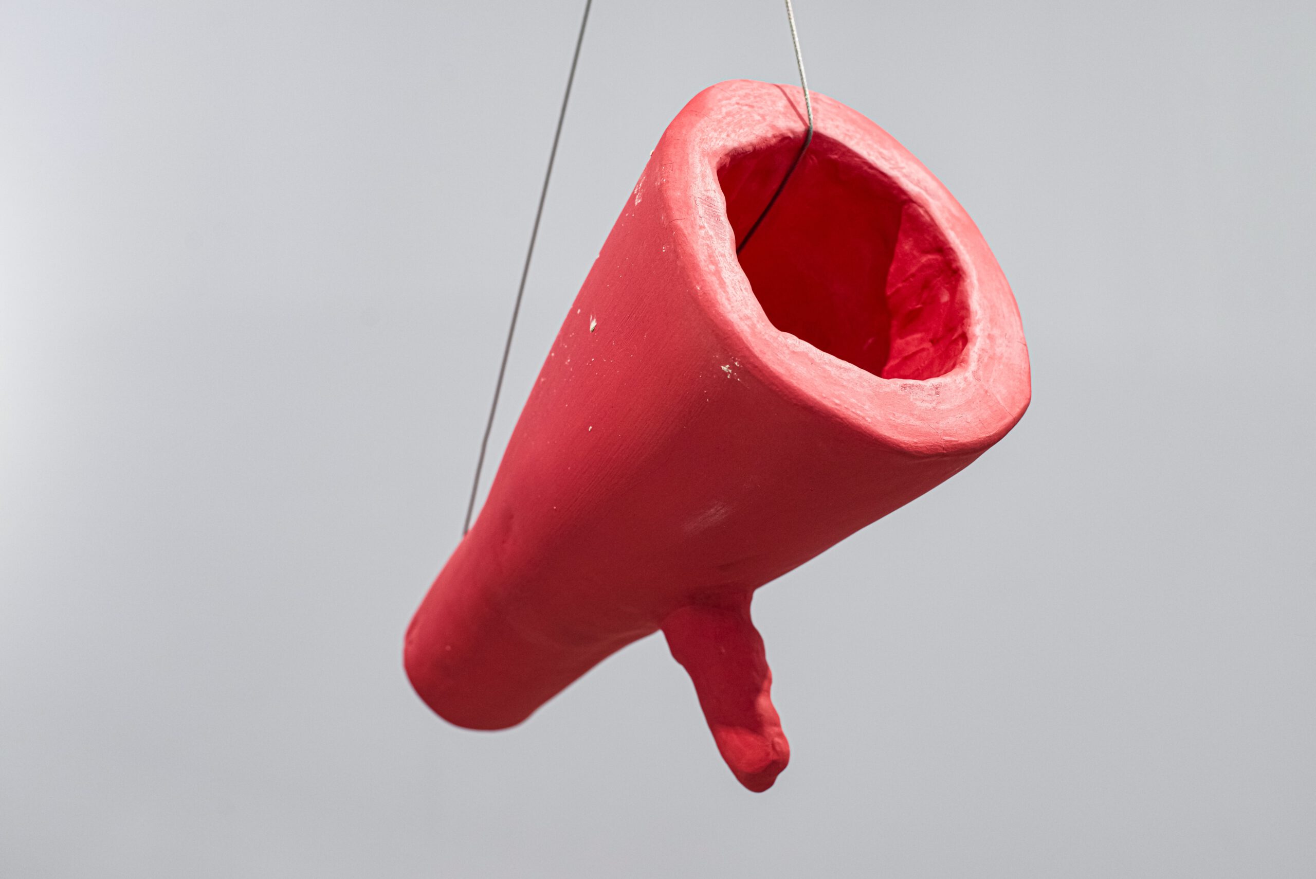 Sculpture by artist Laura Mahnke: Bright red ceramic megaphone hanging from the ceiling in front of a gray wall.