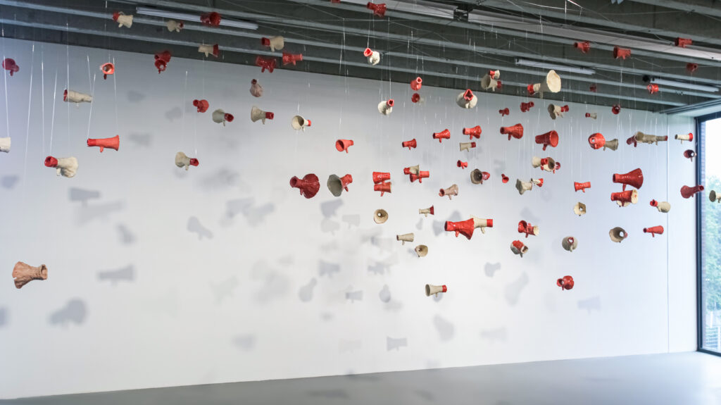 Sculptural installation by artist Laura Mahnke. 80 red ceramic megaphones hanging from a high concrete ceiling.