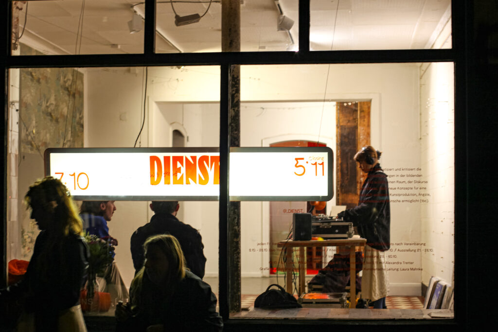 atmospheric night view from the outside into an illuminated gallery window with people and a DJ